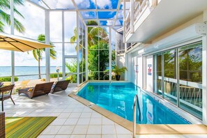 A pool area with a view of the ocean, a covered patio with chairs, and a glass-enclosed structure. - Pools of the Kai (North Side)