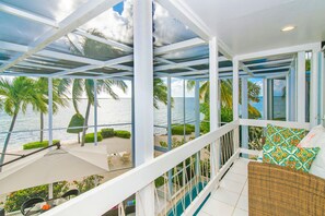 A balcony with a view of the sea, a wicker sofa with patterned cushions, and a glass table with a bottle. - Pools of the Kai (North Side)