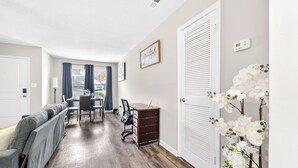 A room with a wooden floor, a grey sofa, a desk with drawers, a chair, a dining table with chairs, a window with curtains, and a wall-mounted thermostat. - Charming 1-bedroom apartment with AC in vibrant Durham (Durham)