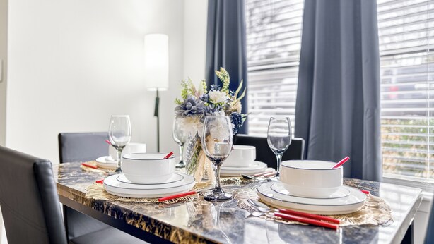 A dining table set with white plates and glasses, a vase with flowers, and a lamp in a room with a window and curtains. - Charming 1-bedroom apartment with AC in vibrant Durham (Durham)