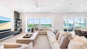 Ein modernes Wohnzimmer mit einer großen Couch, einem Couchtisch und einem Fernseher an der Wand. Große Fenster bieten Blick ins Freie.