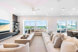 A modern living room with a large sofa, a coffee table, and a TV on the wall. There are large windows with a view of the outdoors. - Aqua 2 on Seven Mile Beach (Seven Mile Beach)