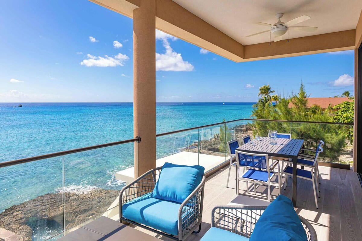 Ein Balkon mit Tisch und Stühlen, der Blick auf das klare, blaue Meer und die felsige Küste freigibt.