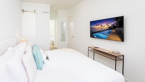 Une chambre à coucher avec un lit, des oreillers, une table de chevet avec une télécommande et un téléviseur fixé au mur qui diffuse une image de coucher de soleil sur une piscine.