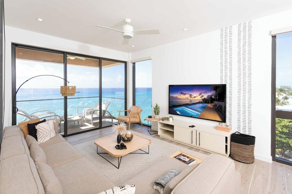 Ein modernes Wohnzimmer mit großem Fernseher, bequemen Sitzmöglichkeiten, einem Couchtisch und Blick auf den Ozean.