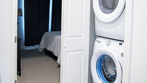 Een wasruimte met twee gestapelde wasmachines, een bed met een witte dekbedovertrek en een donker gordijn op de achtergrond.