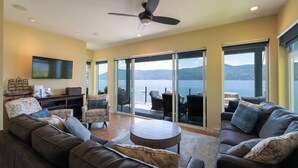 A spacious living room with a large sofa, a coffee table, and a flat-screen TV mounted on the wall. There is a ceiling fan, a fireplace, and a view of the outdoors through large sliding glass doors. - Private Dock, Kayaks, SUPs, Lakefront Views (Celista)