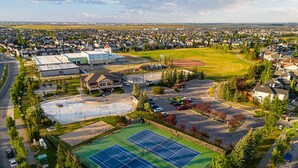 Vue aérienne d’un quartier résidentiel comprenant un grand complexe sportif, avec notamment des courts de tennis et un terrain de football, le tout entouré de maisons et d’un immeuble commercial.