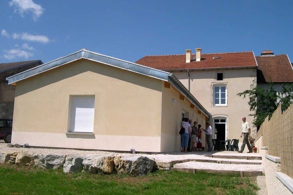 A two-story building with a beige facade and a red-tiled roof. There is a small window on the ground floor and a larger window on the upper floor. A group of people is gathered near the entrance, and a man is walking towards the building.