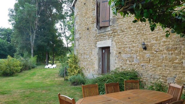 A stone building with a wooden table and chairs in the foreground, surrounded by a grassy garden and trees. - The Récourt Gardens (Récourt)