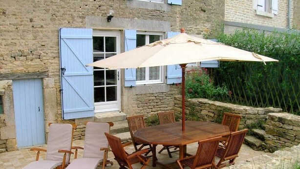 Une terrasse extérieure avec une table et des chaises en bois, un parasol beige et un mur en pierre.