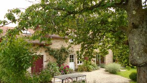 Un bâtiment en pierre avec une porte rouge, entouré d’un jardin avec un chemin de gravier et des meubles d’extérieur.