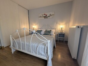 Un dormitorio con una estructura de cama metálica blanca, una mesita de noche con lámpara, un radiador y una obra de arte en la pared.