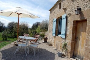Un'area pranzo all'aperto con tavolo e sedie, un ombrellone bianco e un edificio in pietra con persiane blu.