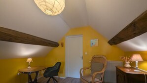 A room with a sloped ceiling, a table with a lamp, a chair, a wicker chair, and a radiator. - Simone de Beauvoir Guesthouse (Châteauvillain)
