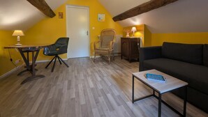 A room with a wooden ceiling, a black sofa, a small table, a chair, and a lamp. - Simone de Beauvoir Guesthouse (Châteauvillain)