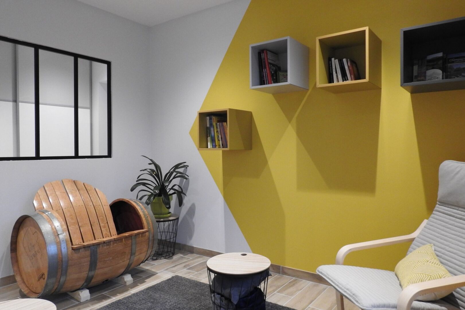 A modern living room with a yellow accent wall, a wooden barrel seating area, a round table, and a white armchair with a cushion.