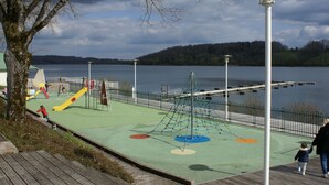 Un parc infantil amb tobogans i elements per escalar a prop d'un llac, envoltat per una tanca i un fanal.