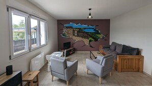 Un ampli saló amb una finestra gran, televisió de pantalla plana, butaques grises, un sofà de fusta i un mural a la paret.