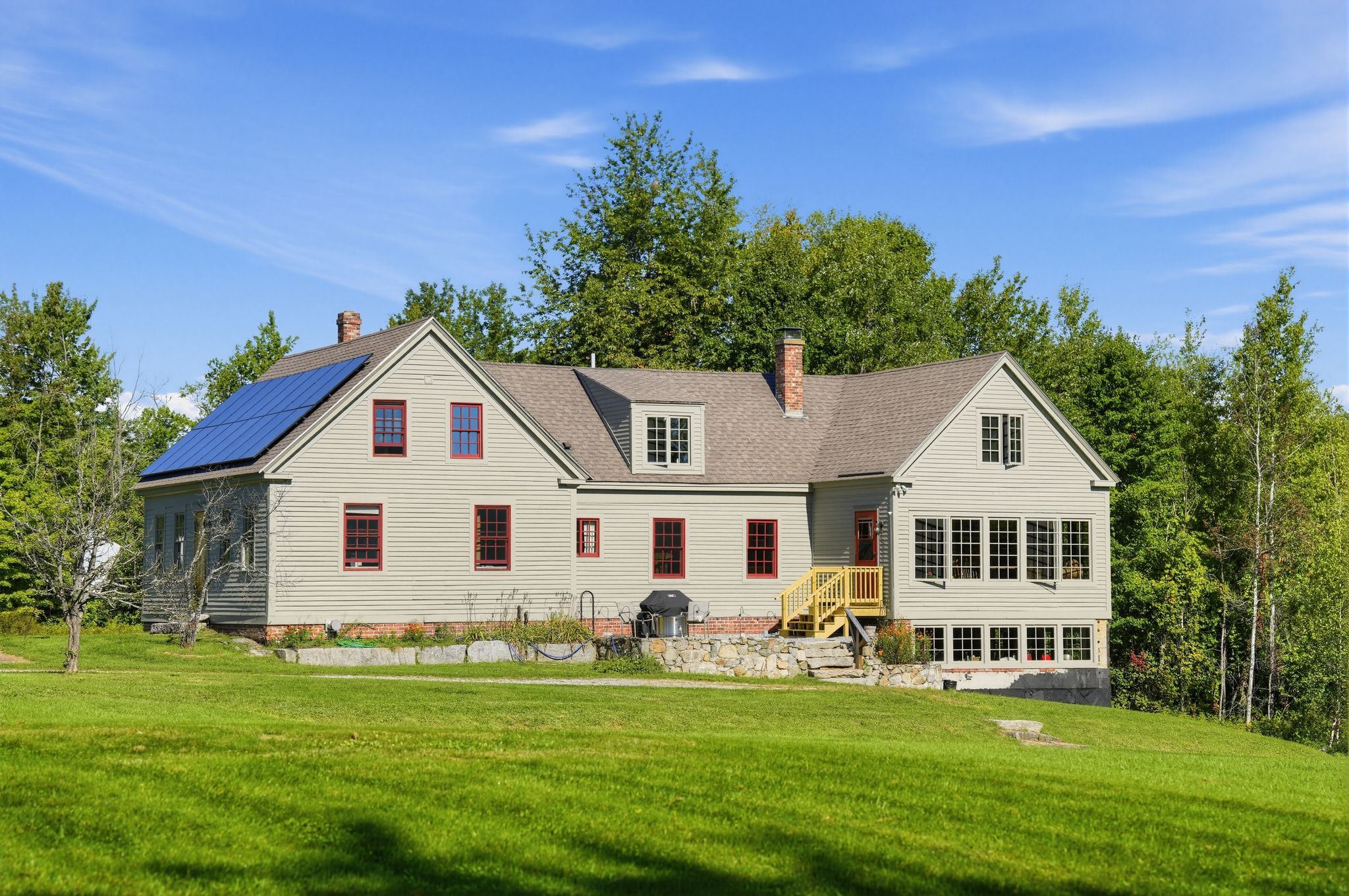 Una casa de dos pisos con un amplio porche delantero, paneles solares y cimientos de piedra.