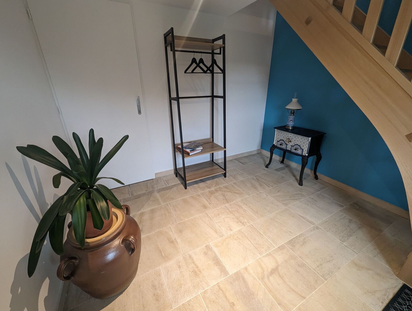 Ein Zimmer mit Fliesenboden, einer Topfpflanze, einem Metallregal und einem kleinen Tisch mit Lampe.