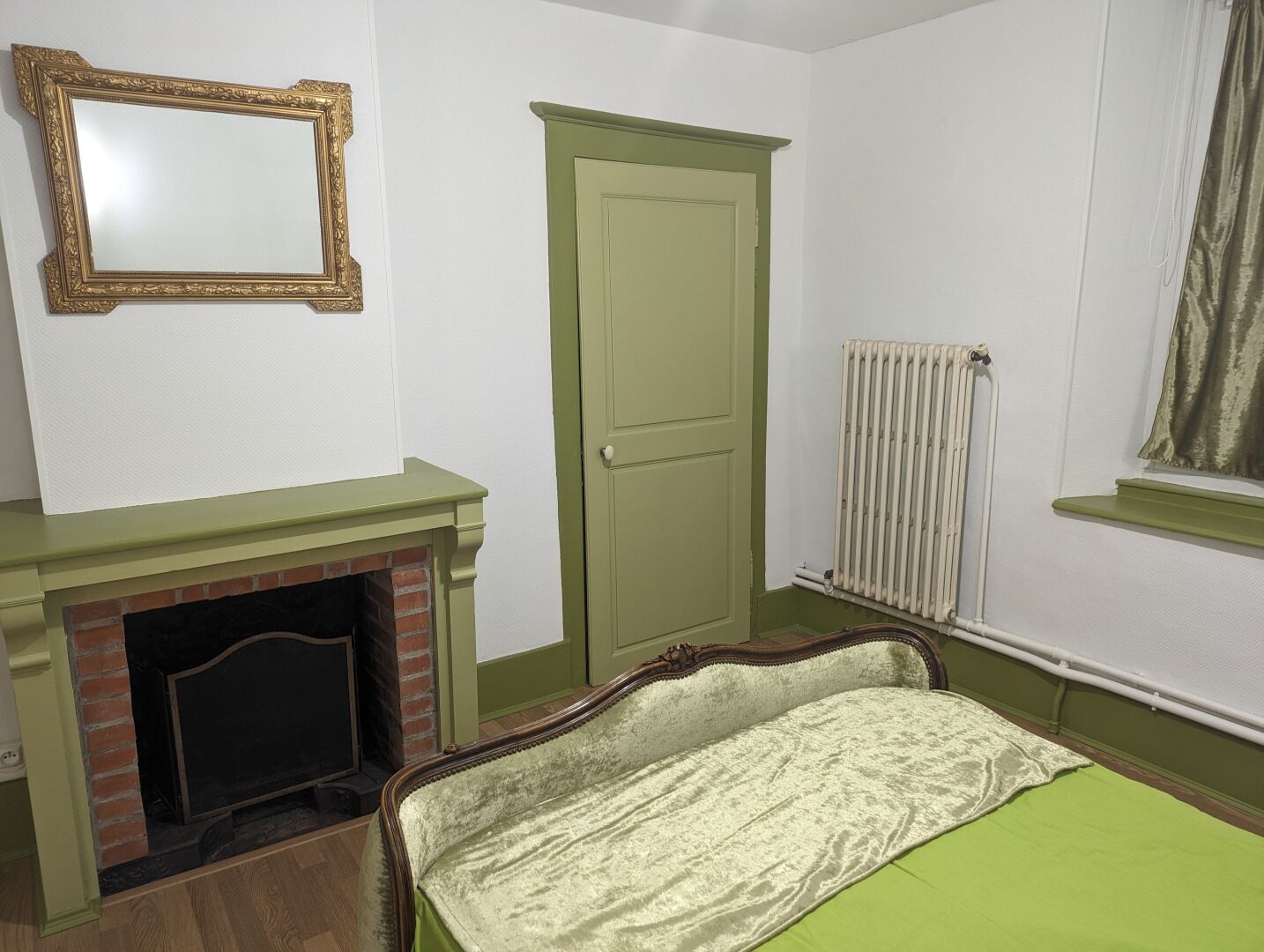 A room with a green door, a fireplace, a radiator, and a bed with a green bedspread.