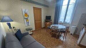 Una sala de estar con un sofá, una mesita de noche con una lámpara, un cuadro en la pared, un comedor con una mesa y sillas, un televisor y una ventana con cortinas.
