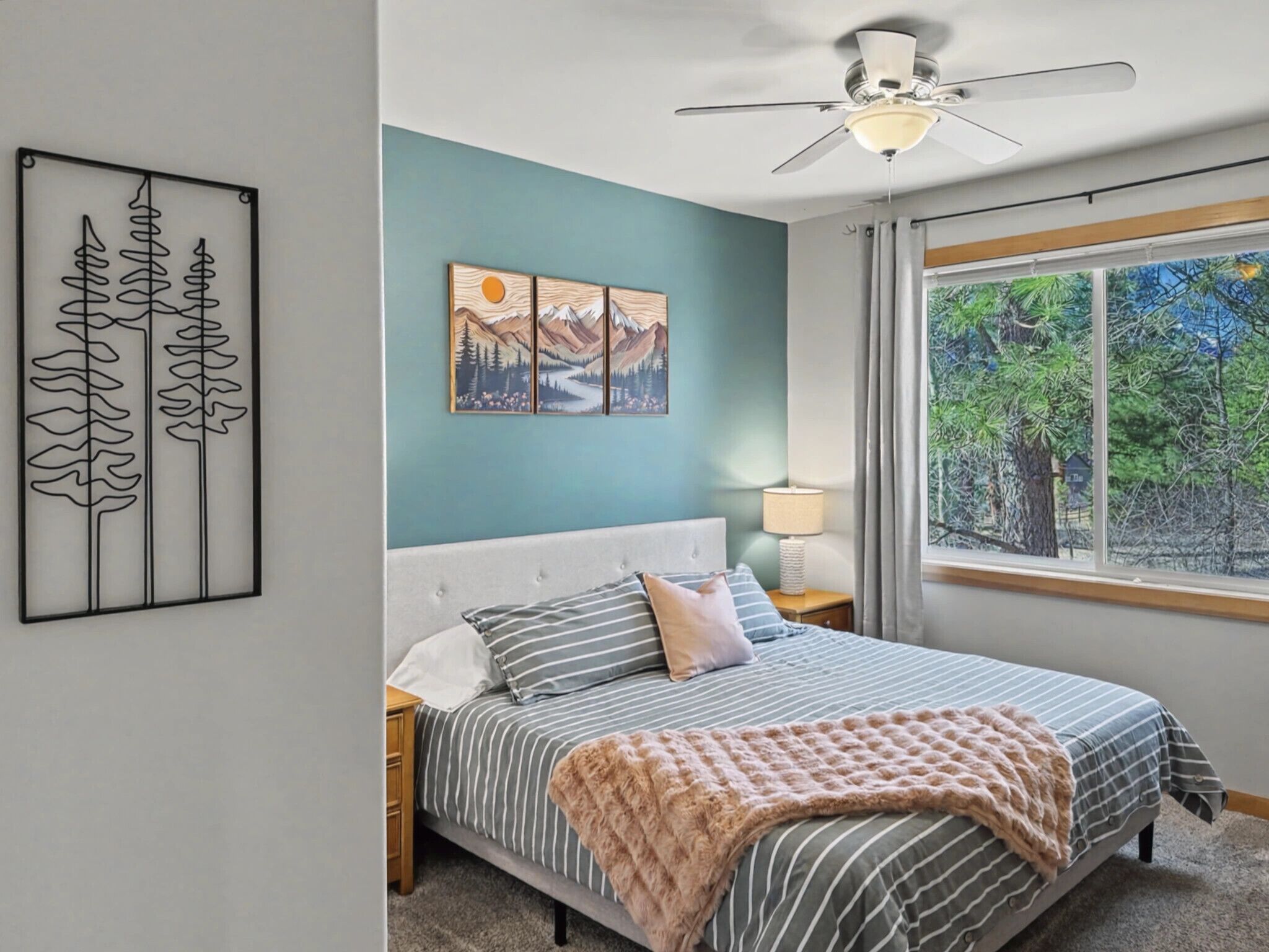 A bedroom with a bed, a ceiling fan, a window with a view of trees, and a wall with a painting.