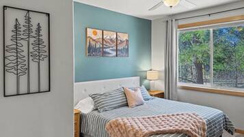 A bedroom with a bed, a ceiling fan, a window with a view of trees, and a wall with a painting.