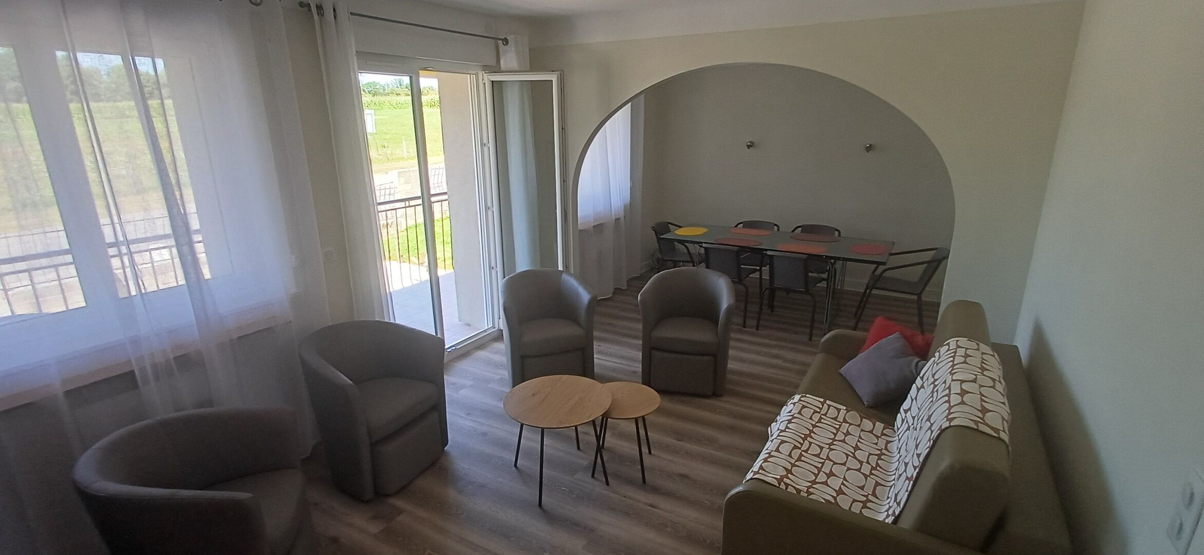 Een woonkamer met een bank, twee fauteuils, een salontafel en een eettafel met stoelen. Er is een schuifdeur die leidt naar een buitenruimte.