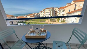 Un balcone con un tavolo apparecchiato per due, con due bicchieri di vino e un vassoio con una candela. Il balcone si affaccia su una zona residenziale con edifici e vista sul mare.
