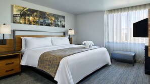 Habitación de hotel con una cama grande, dos mesitas de noche, un televisor de pantalla plana, una ventana con cortinas translúcidas y un cuadro de paisaje urbano en la pared.