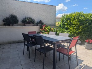 Eine Terrasse im Freien mit einem rechteckigen Tisch und sechs Stühlen, umgeben von Topfpflanzen und einem großen Strauch.