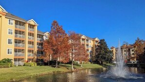 Un immeuble résidentiel à plusieurs étages doté de balcons, un étang avec une fontaine et des arbres aux couleurs automnales.