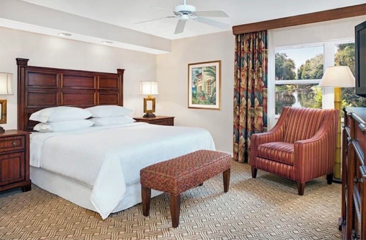Quarto de hotel com uma cama grande, cabeceira de madeira, um banco, uma cadeira, mesa de cabeceira, abajur, pintura e janela com cortinas.