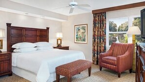 Une chambre d’hôtel avec un grand lit, une tête de lit en bois, un banc, une chaise, une table de chevet, une lampe, un tableau et une fenêtre avec des rideaux.