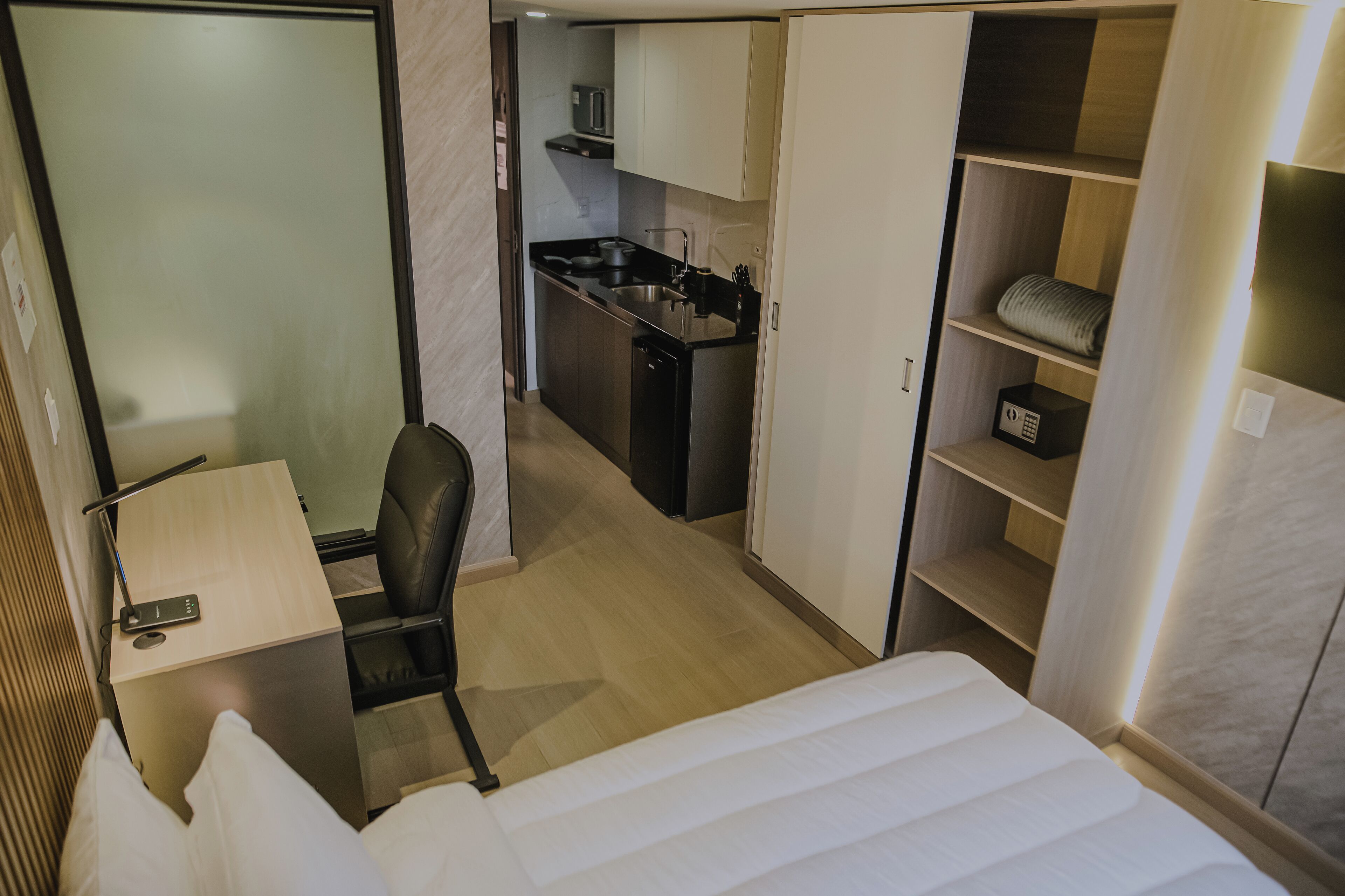 Quarto de hotel com cama, escrivaninha, cadeira, cozinha compacta e guarda-roupa.