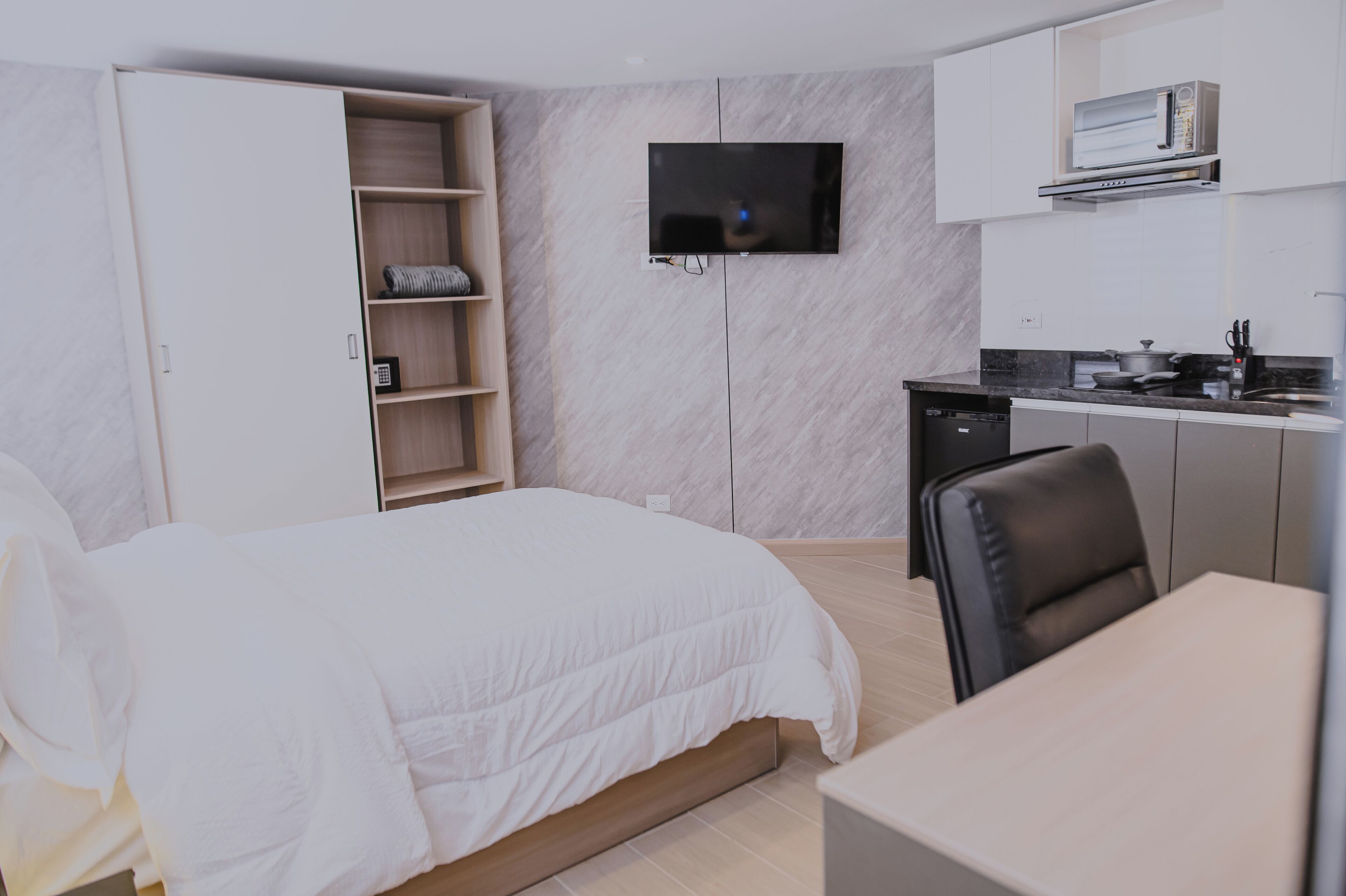 A hotel room with a bed, a TV mounted on the wall, a kitchen area with a microwave, and a dining table.