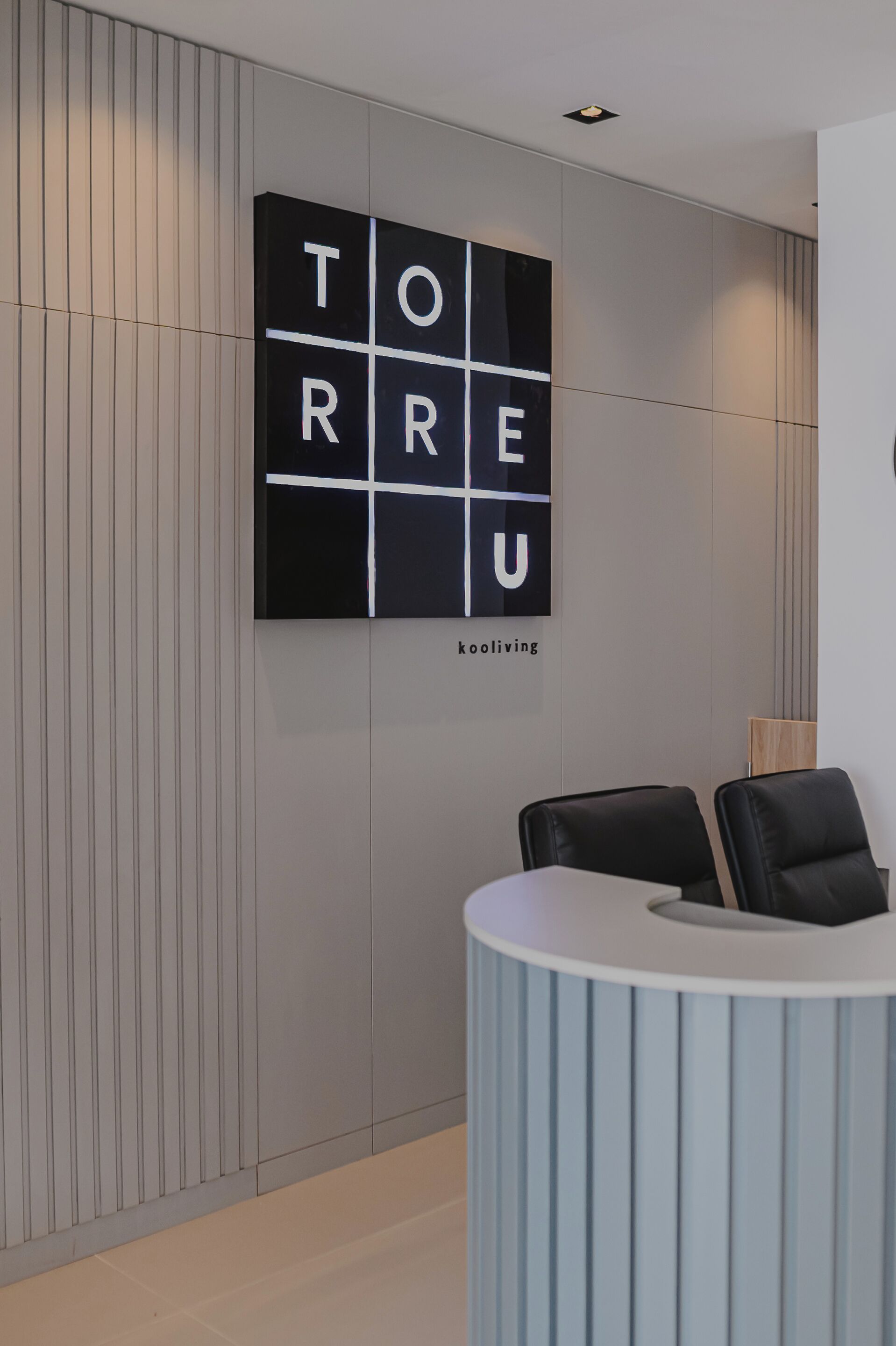 Een receptiegedeelte met een modern design, met een wandbord waarop de naam 'TORRE' en een logo staan. Er zijn twee zwarte stoelen en een wit receptiebalie.