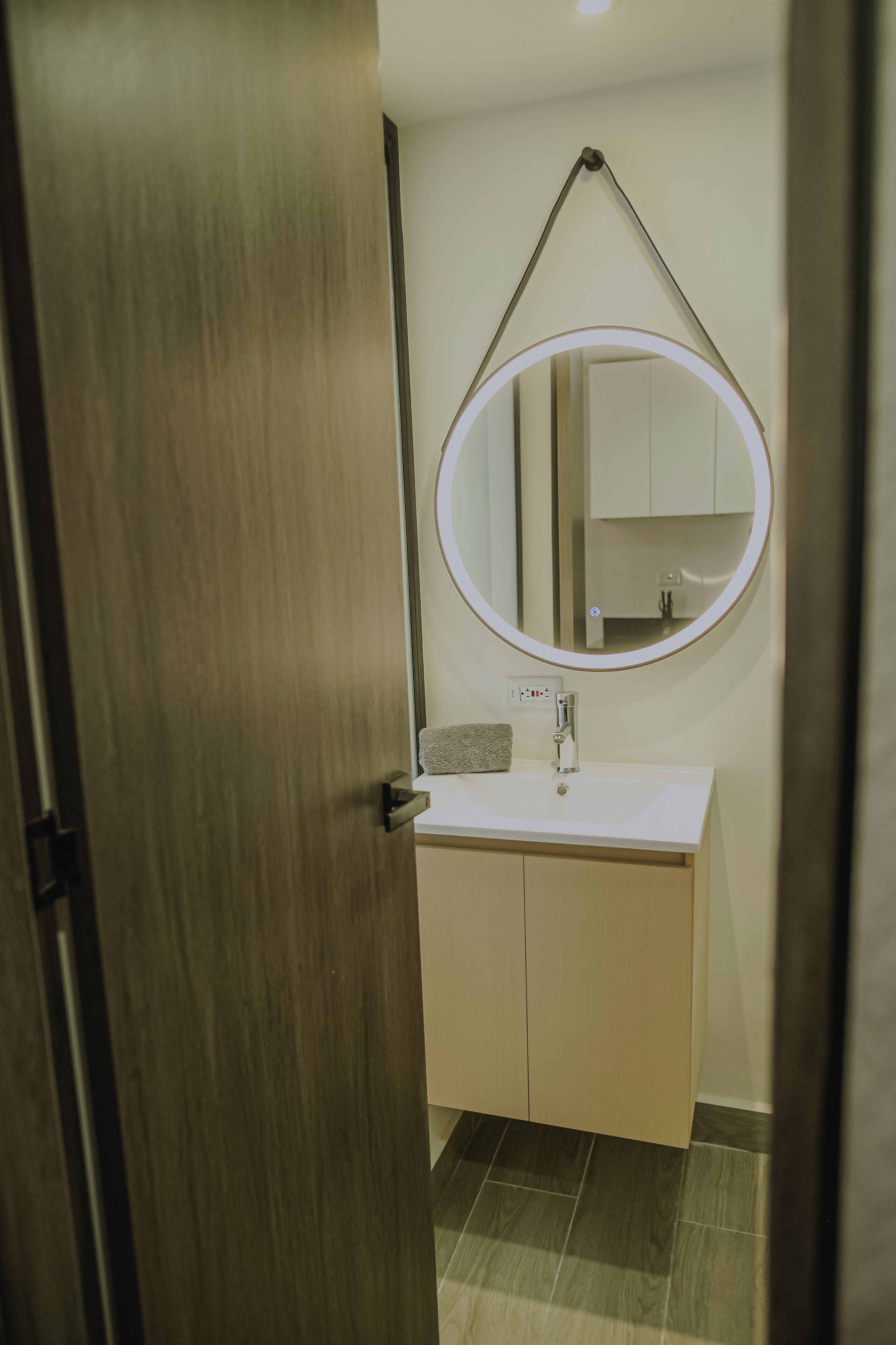 Un bany amb una porta de fusta, un lavabo blanc, un mirall amb llum i un sòl de rajola.
