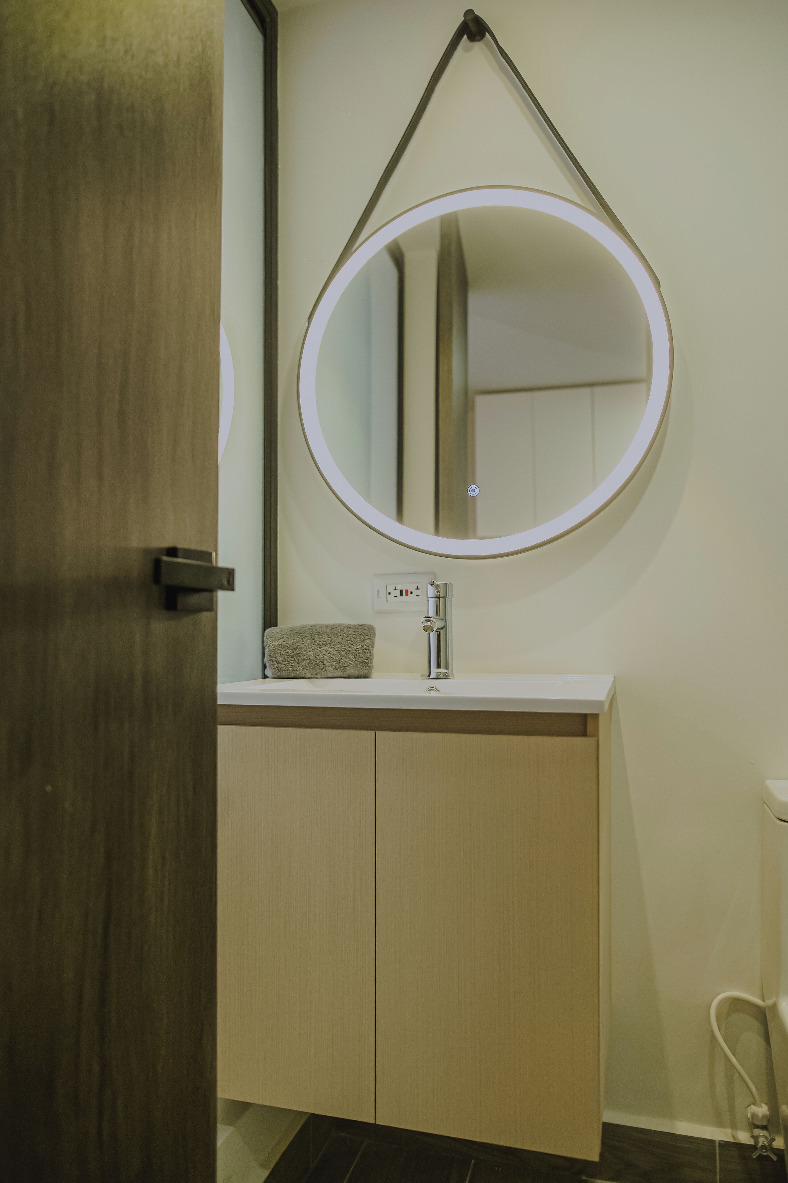 A bathroom with a wooden door, a white sink with a towel, and a round mirror with a light.