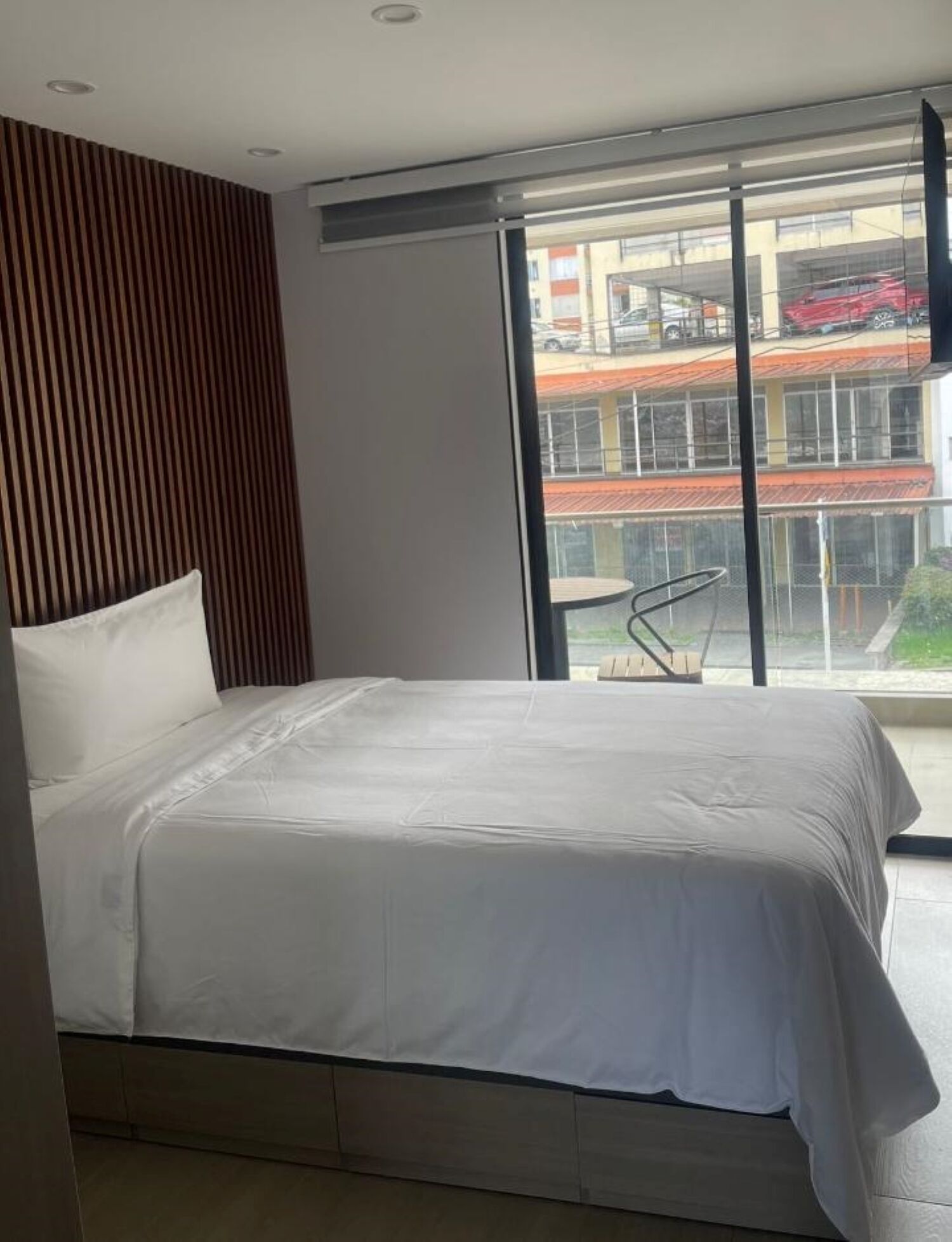 A modern bedroom with a large bed, a wooden slat wall, and a view of a building with a red car.