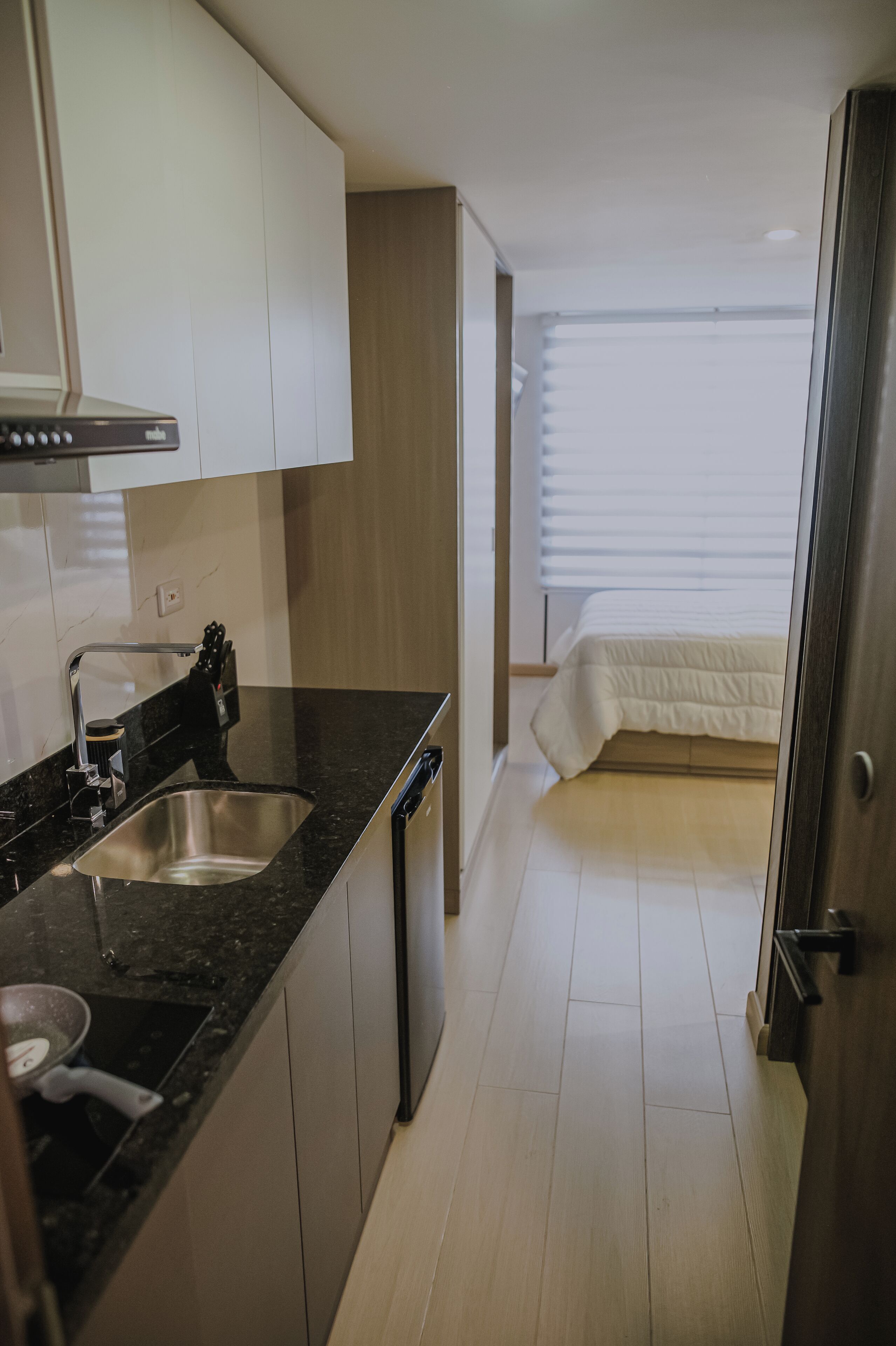 A compact kitchen with a sink, stove, and cabinets, leading to a bedroom with a bed and window blinds.