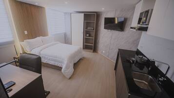 Una habitación de hotel moderna con cama, escritorio, televisor montado en la pared y una zona de cocina con lavabo y estufa.