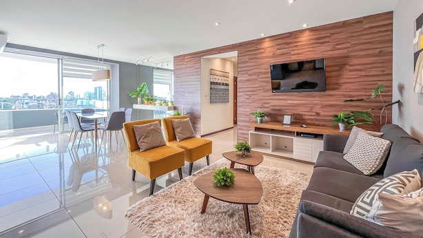 Uma sala moderna com sofá, mesa de centro e cadeiras, um espaço de jantar com mesa e cadeiras, e vista para a cidade através de amplas janelas.