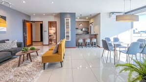 Uma sala moderna com sofá, banqueta e mesa de centro, uma área de jantar com cadeiras e uma cozinha com balcão.