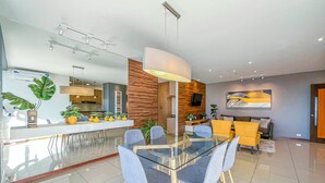 Uma sala de jantar moderna com mesa de vidro e cadeiras azuis, uma parede de destaque em madeira e uma cozinha ao fundo.