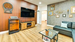 A modern living room with a flat-screen TV, wooden panel walls, a sofa, a glass coffee table, and a dartboard. - Downtown Cornelius Bungalow  Walk to Dining & Cain Center Karaoke + Darts (Cornelius)