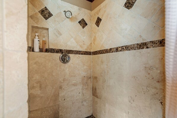 A shower area with beige tiles, a showerhead, and a shelf with toiletries. - Lake Travis Retreat w/ Pool, Hot Tub & Game Room (Lago Vista)