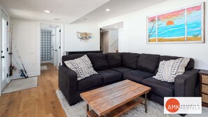 Een moderne woonkamer met een donkere hoekbank, een houten salontafel en een kleurrijk schilderij van een zonsondergang aan de muur.